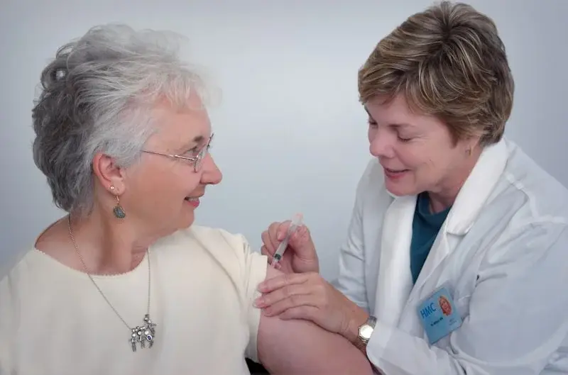 Une soignante en blouse blanche administre un vaccin à une résidente âgée, qui lui sourit.